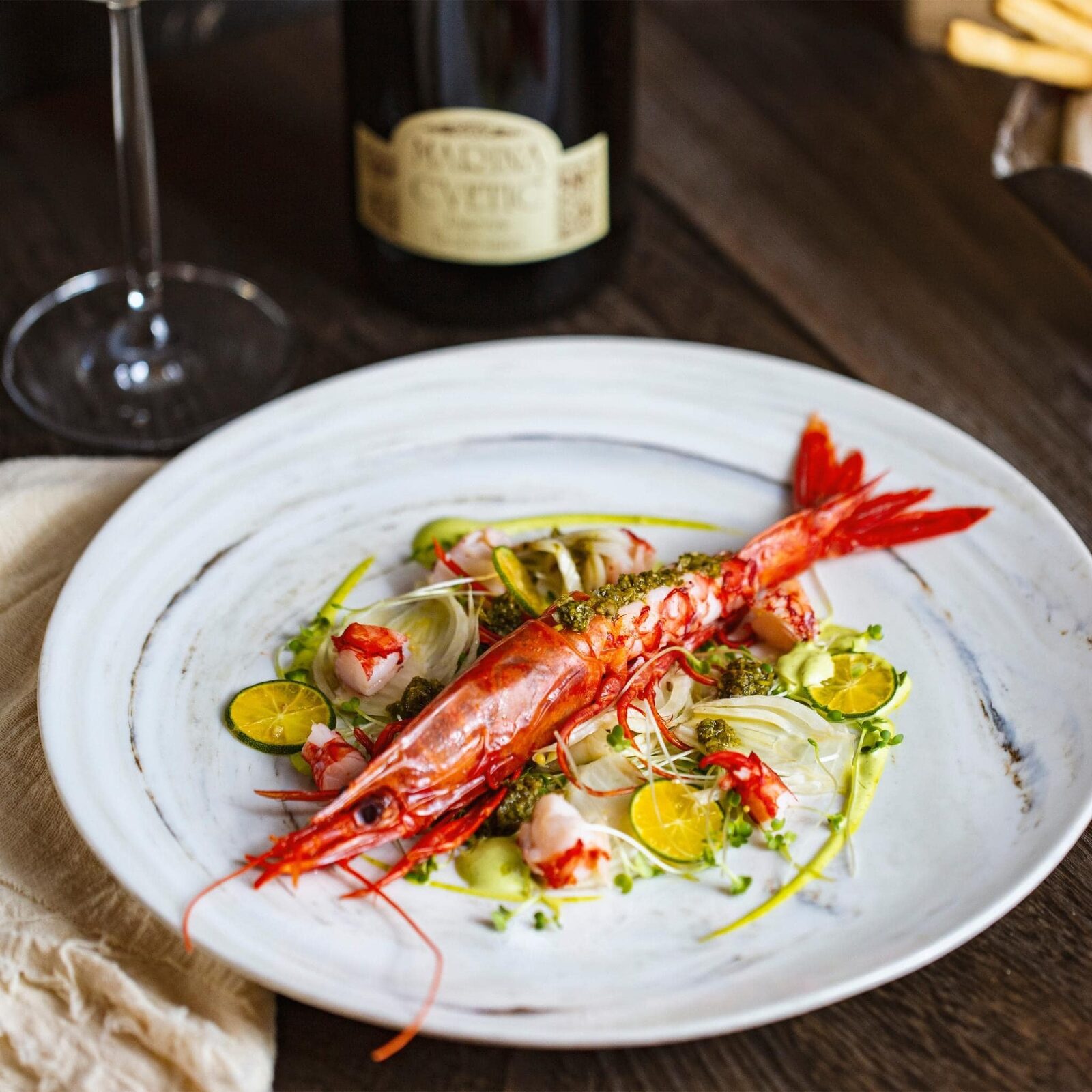 A whole red carabinero prawn on shaved fennel with citrus halves and green herb sauce, a Marina Cvetic wine bottle behind