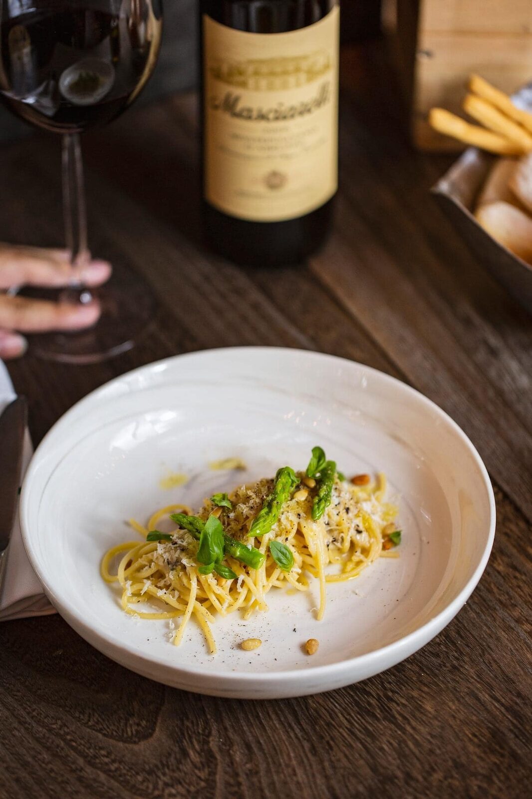 A bowl of spaghetti with asparagus, grated cheese and pine nuts in front of a Masciarelli bottle and a glass of red wine