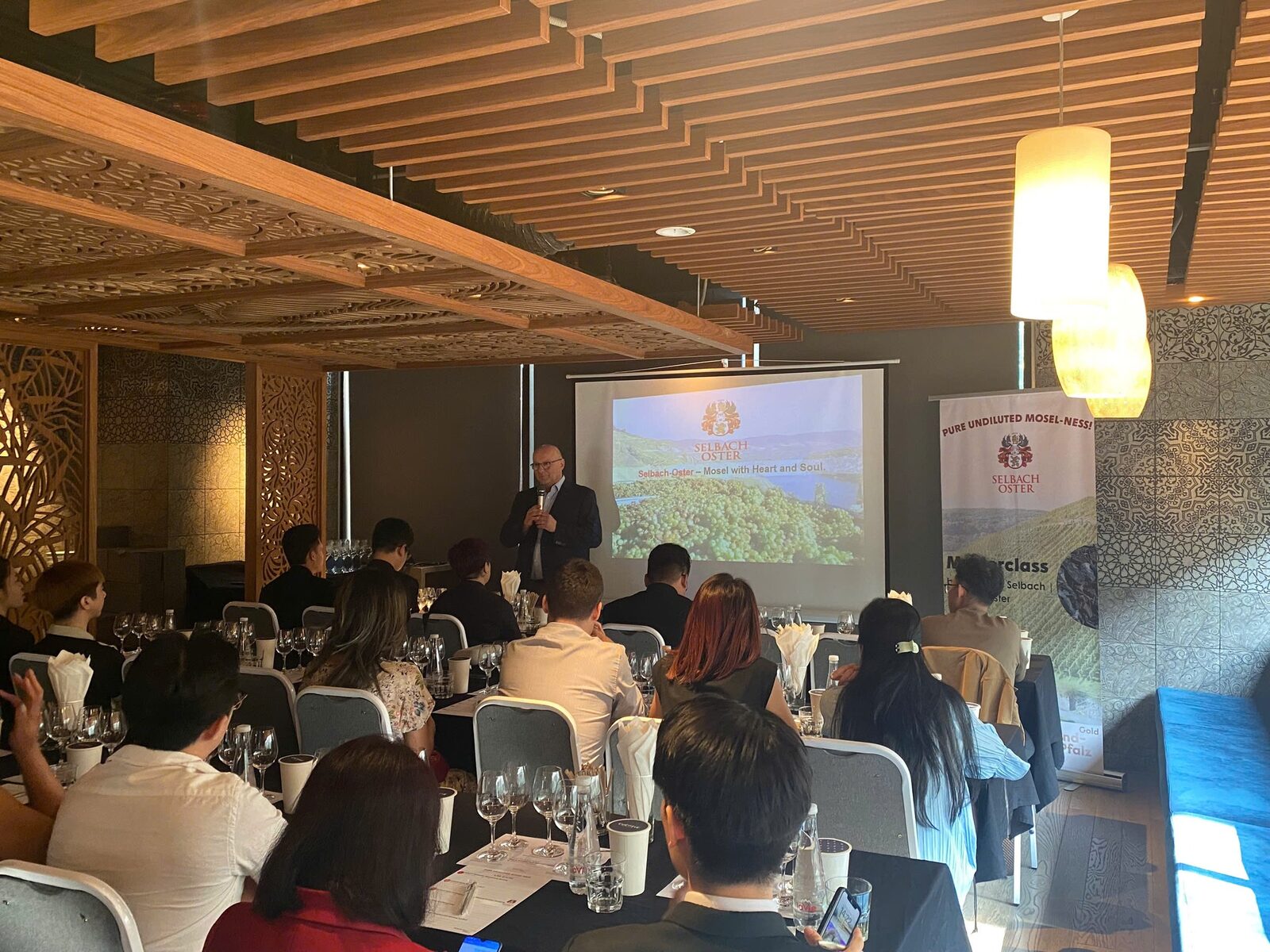 Wide view of the masterclass room with tasting glasses on tables and a speaker in front of a projected vineyard image