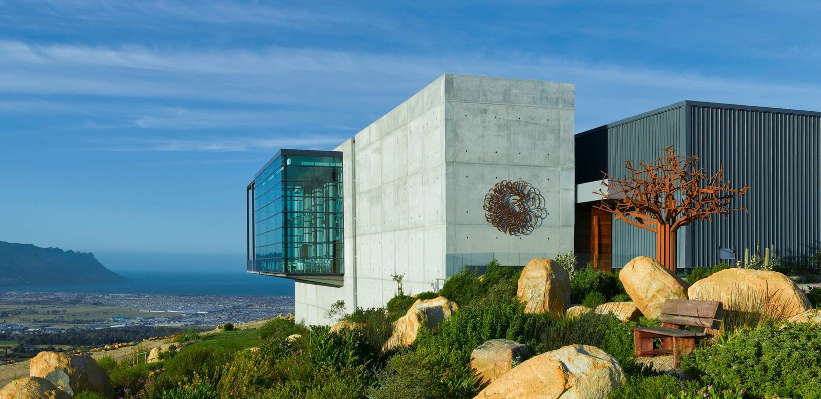 Modern concrete and glass winery building on a hillside with fynbos, boulders and the sea in the distance